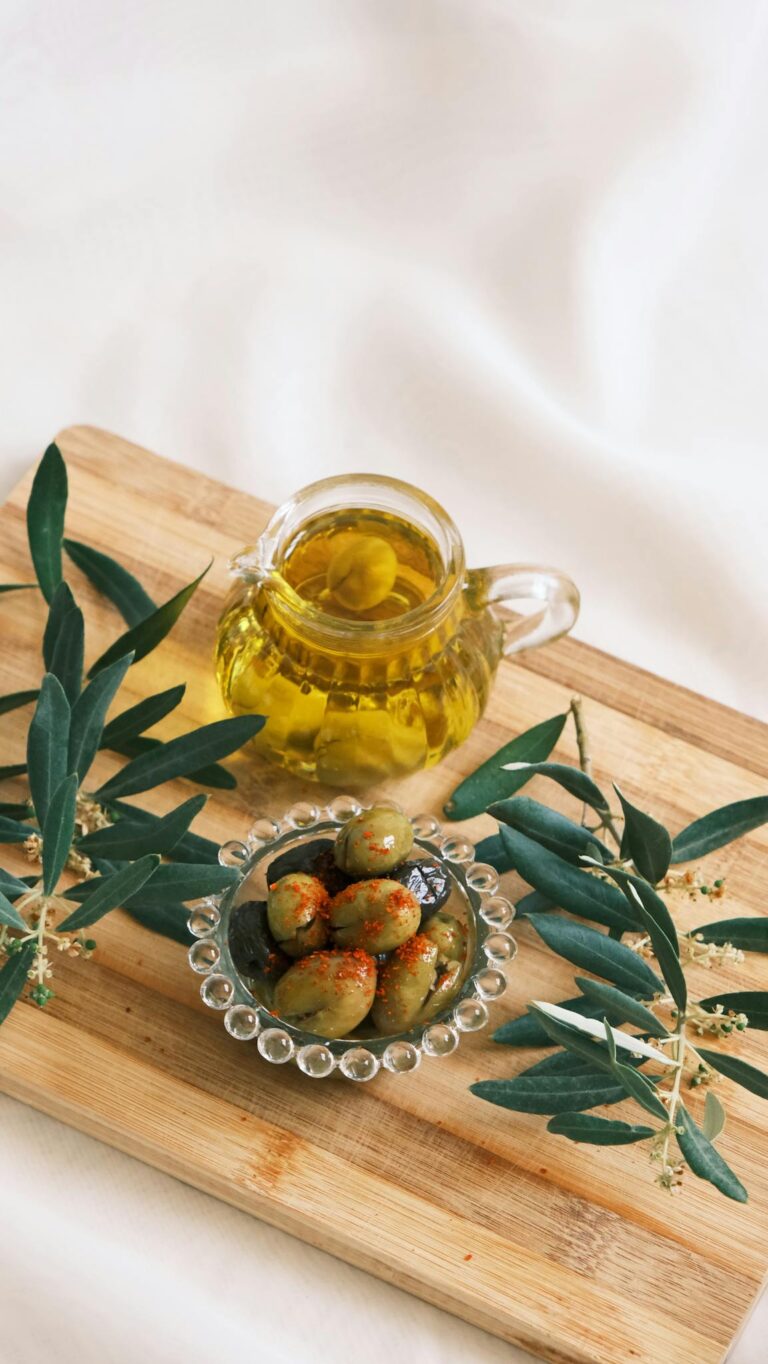 Glass jug of olive oil and a bowl of olives on a wooden cutting board with olive branches.